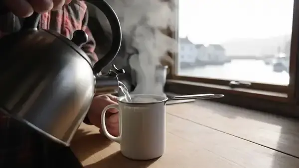 A man in his sixties pouring hot water into a white mug with a metal tea infuser at a kitchen counter